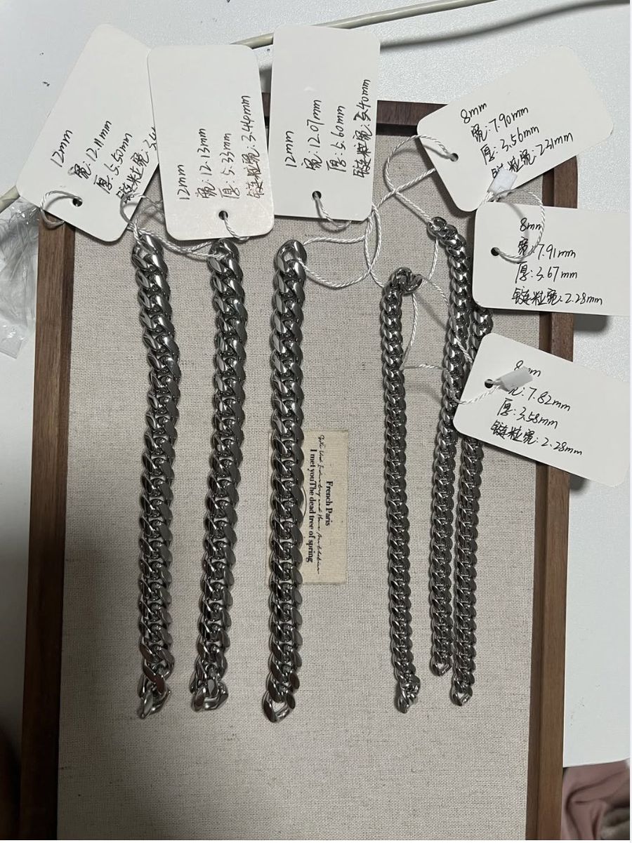 Seven pre-production Cuban link chains hanging on a board, each with a handwritten tag recording its measured width and thickness